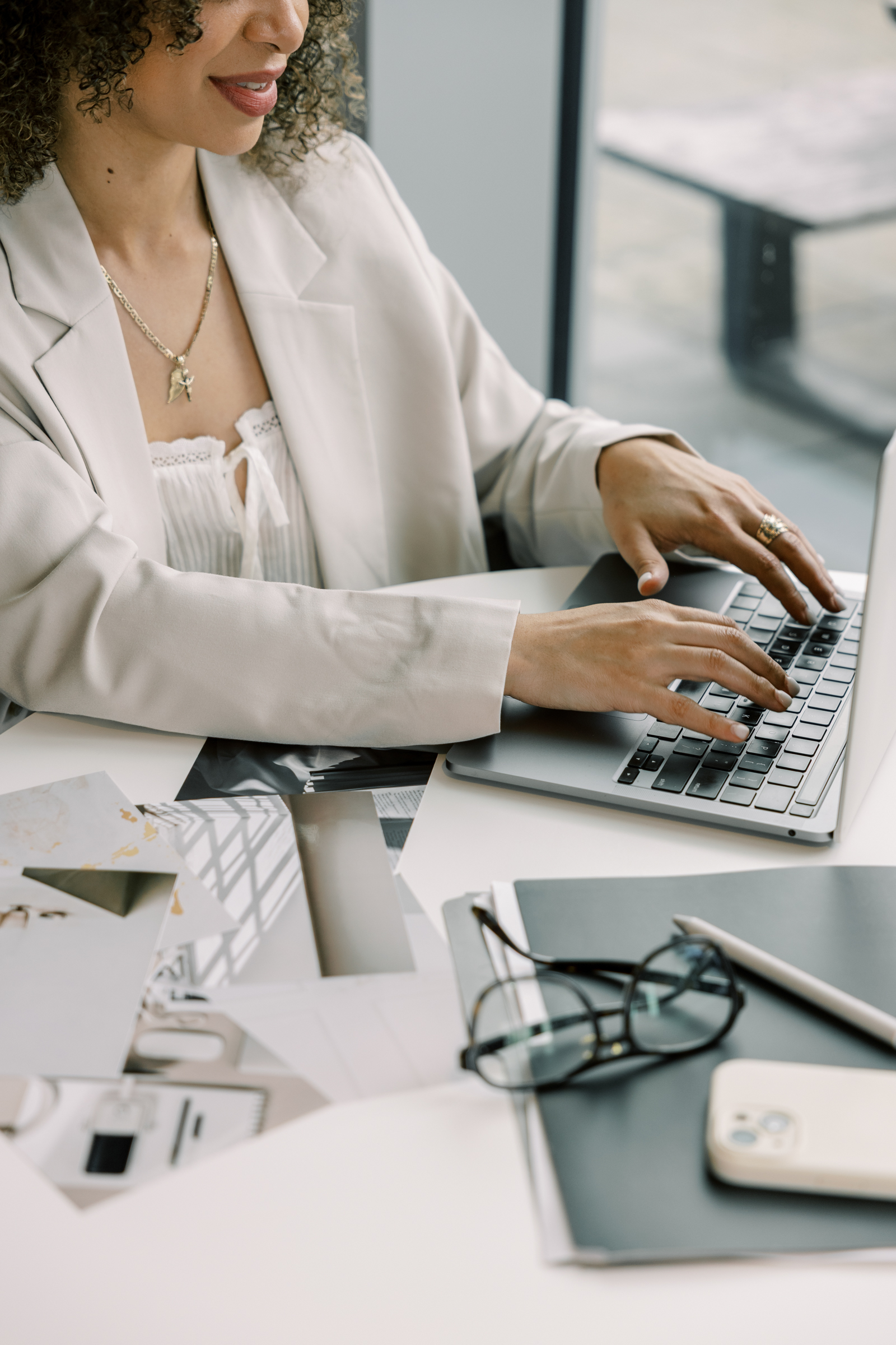 Woman in a white suite is typing on her laptop which is on a desk with other work related items like glasses, a phone, files, and photos.