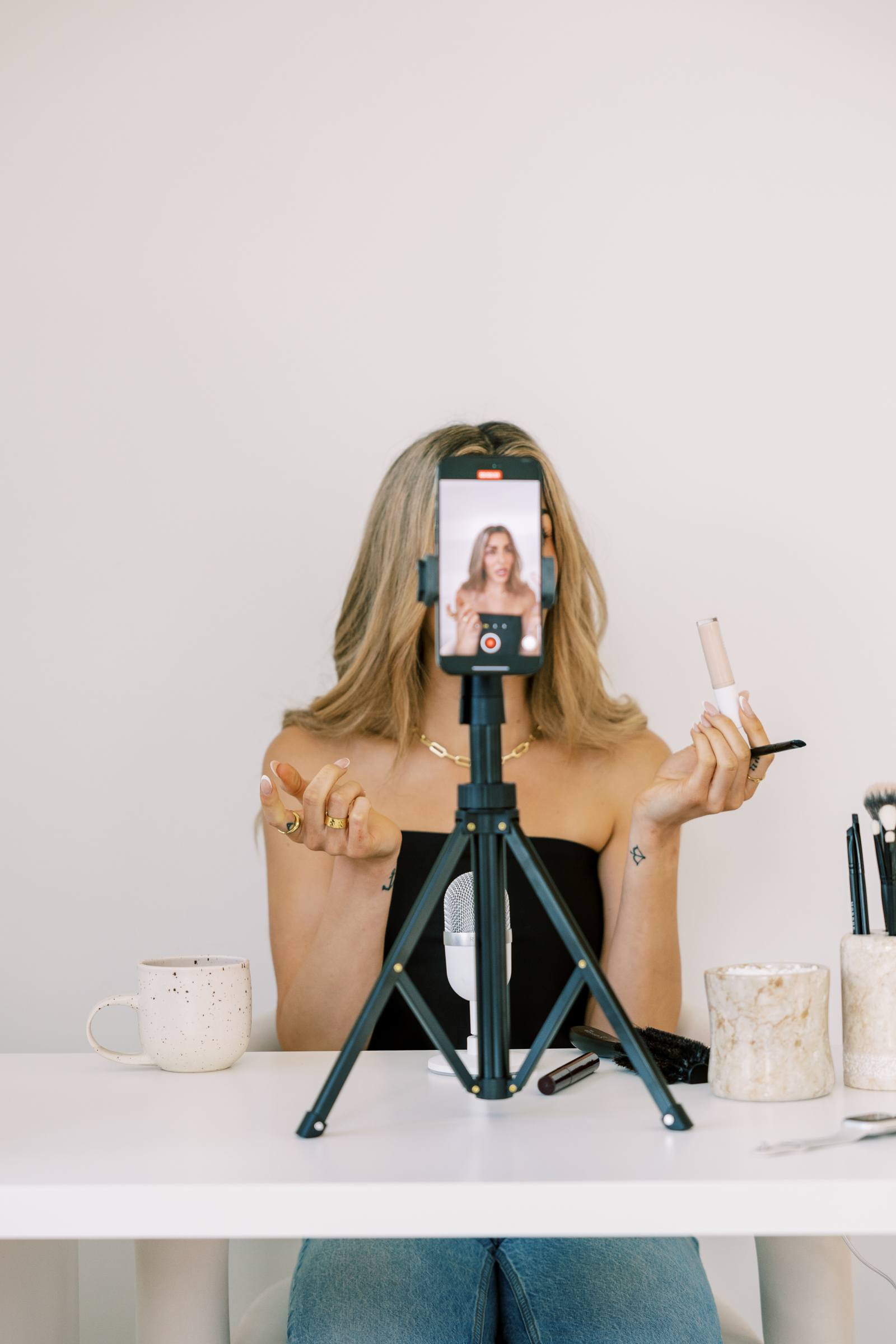 Woman behind a phone camera on a tripod putting on her makeup on a white desk